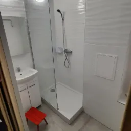 Nowoczesna, jasna łazienka po remoncie z kabiną prysznicową typu walk-in, białą umywalką z szafką, szarymi płytkami podłogowymi i małym czerwonym stołkiem.