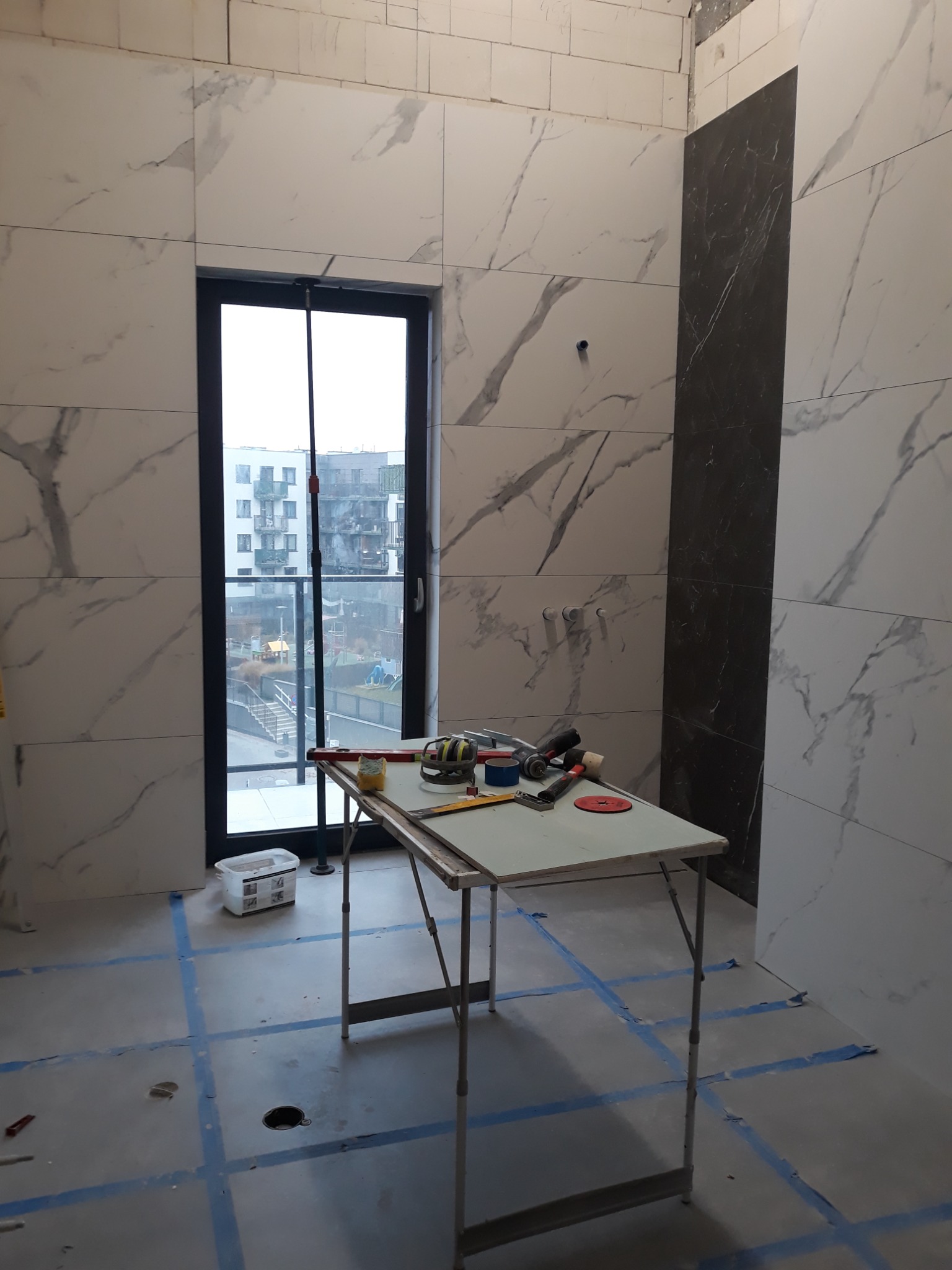 Łazienka w trakcie remontu: białe marmurowe płytki na ścianach, fragment czarnej ściany, okno z widokiem na blok, stół roboczy z narzędziami i akcesoriami glazurniczymi, podłoga z wyznaczonymi...