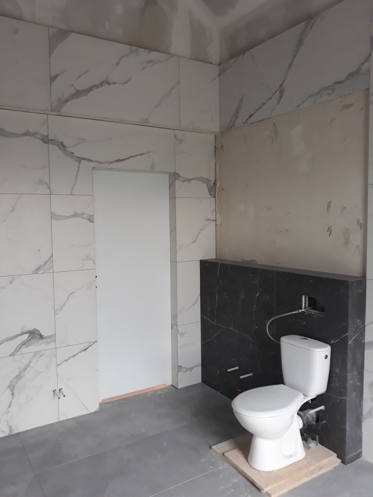 Łazienka w trakcie remontu: białe płytki imitujące marmur na ścianach, szara podłoga, częściowo wykończona ściana z czarnych płytek i zamontowana toaleta na drewnianym podeście.
