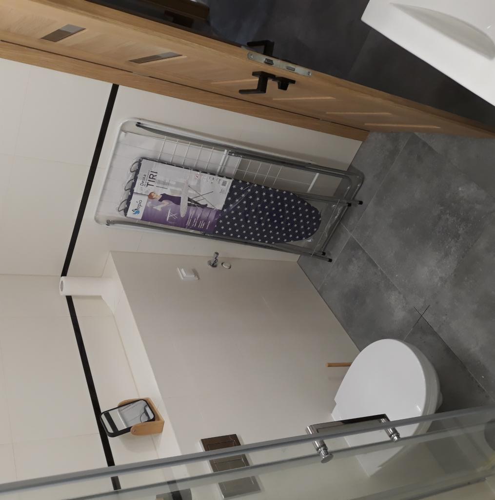 Nowoczesna łazienka z szarymi płytkami podłogowymi, białą toaletą i drewnianymi drzwiami. W rogu stoi rozłożona suszarka na ubrania z deską do prasowania w opakowaniu.