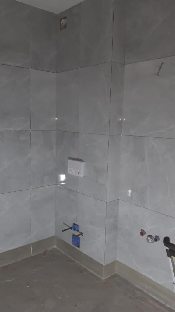 Świeżo wykafelkowana ściana łazienki z szarymi płytkami imitującymi marmur, widoczne wystające rury i puszka elektryczna, surowy beton na podłodze.