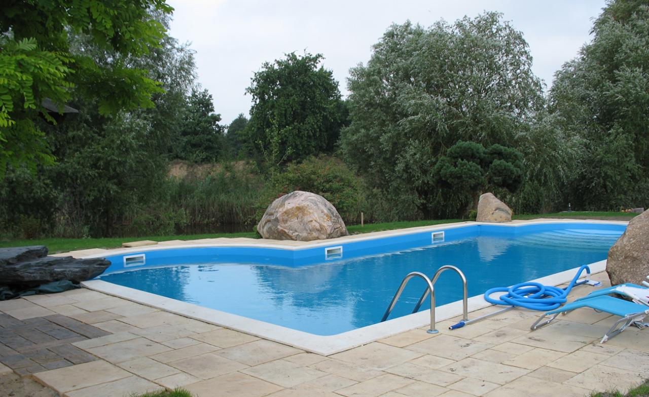 Prostokątny basen w ogrodzie z kamienną obudową i metalową drabinką, otoczony zielenią i dużymi kamieniami dekoracyjnymi, z leżakiem i wężem do czyszczenia na płytkach.