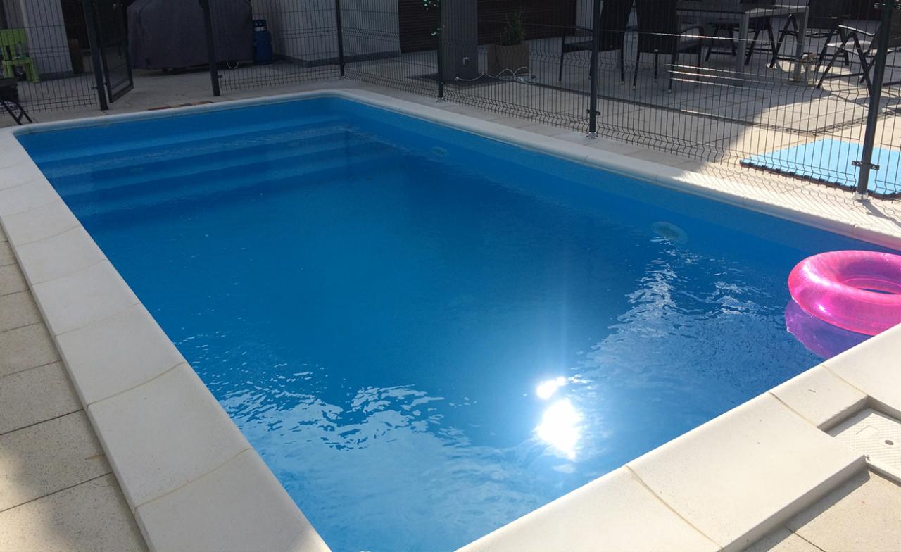 Napełniony, prostokątny basen z jasnoniebieską wodą, z widocznymi schodami wejściowymi i różowym kołem do pływania, otoczony jasnymi płytkami i czarnym metalowym ogrodzeniem.