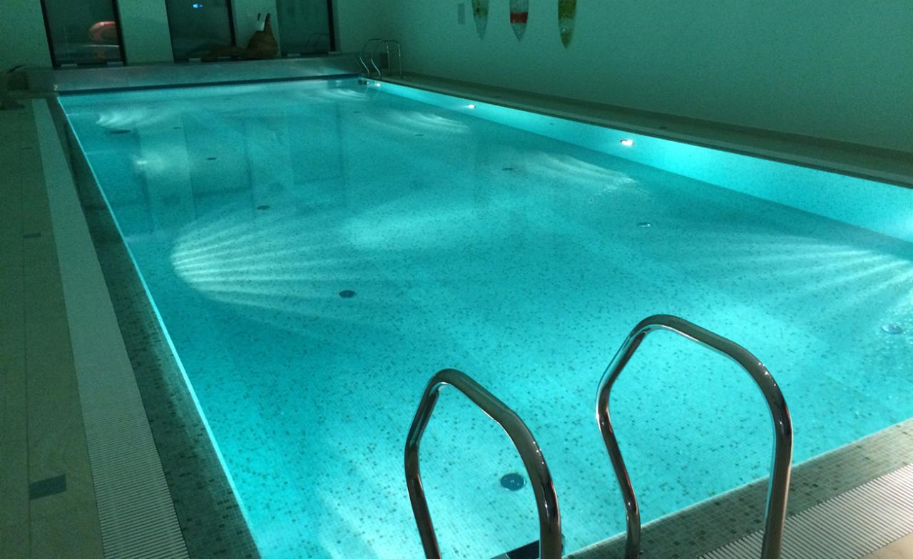 Kryty basen z turkusową wodą, widoczne metalowe schodki wejściowe, oświetlenie punktowe pod wodą tworzące wzór na dnie.
