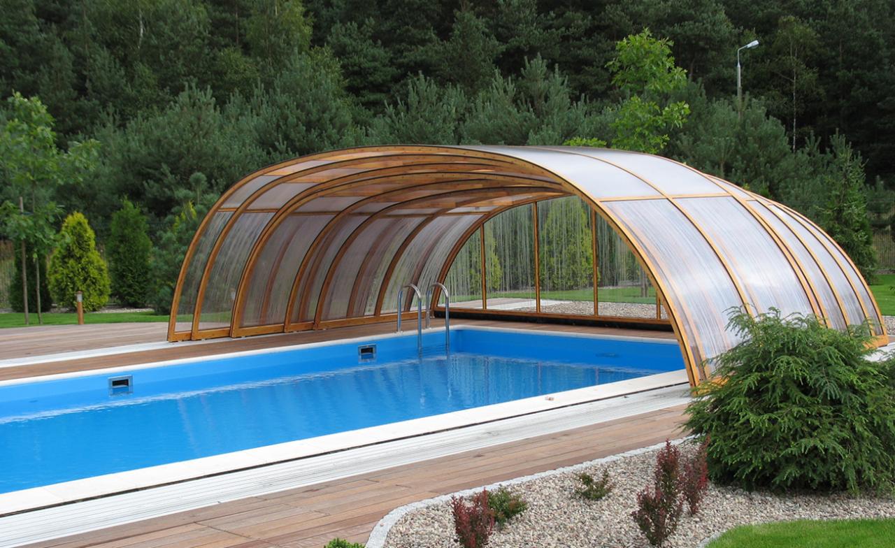 Zadaszony basen ogrodowy z drewnianą konstrukcją łukową i przezroczystym pokryciem, otoczony drewnianym tarasem i zielenią