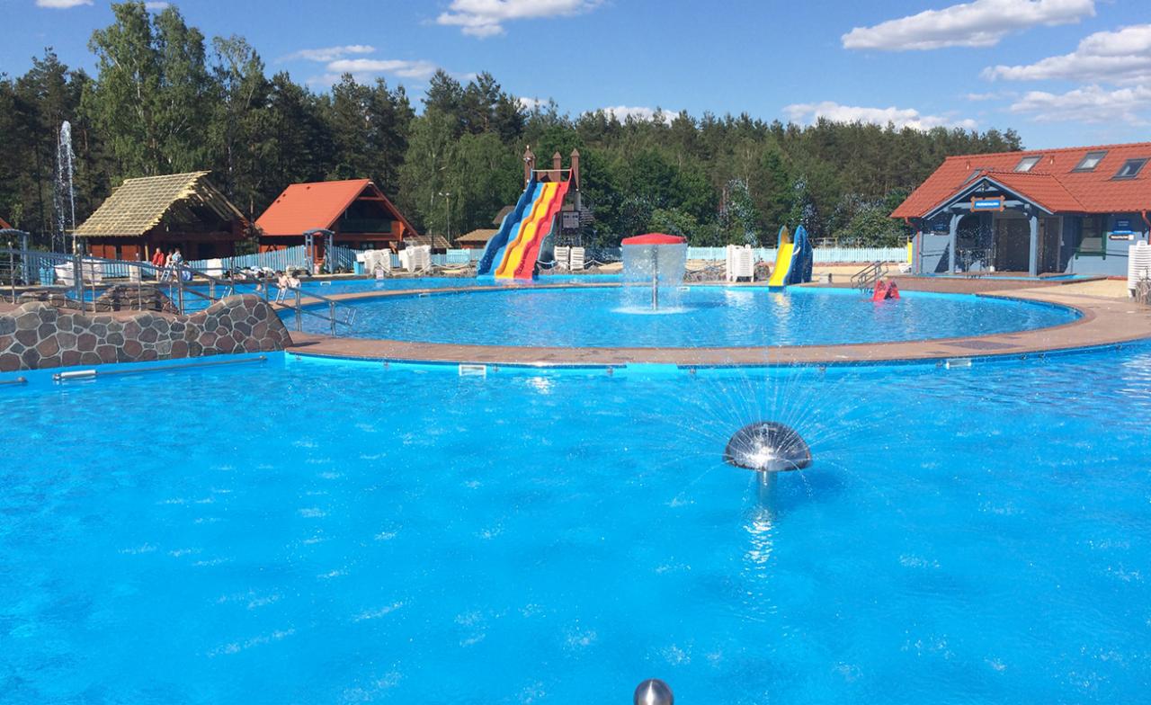 Rozległy, publiczny kompleks rekreacyjny z basenem w Toruniu, wyposażony w zjeżdżalnie, fontanny i drewniane budynki w tle.