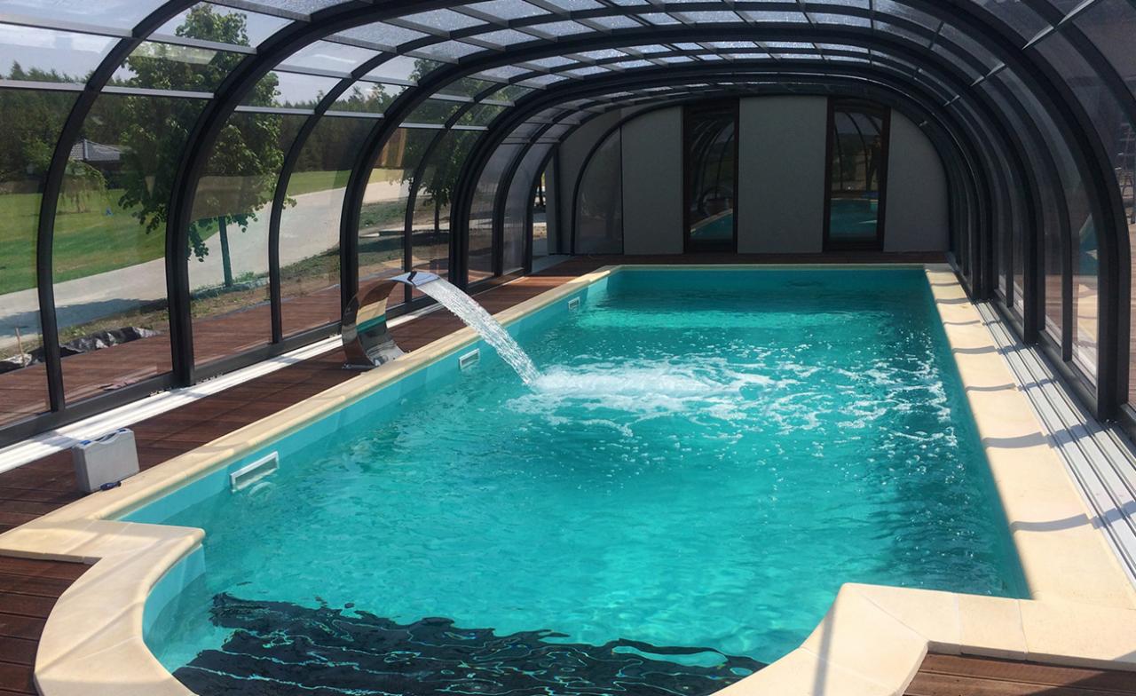 Zadaszony basen z turkusową wodą i stalową kaskadą, otoczony drewnianym tarasem i ogrodem.