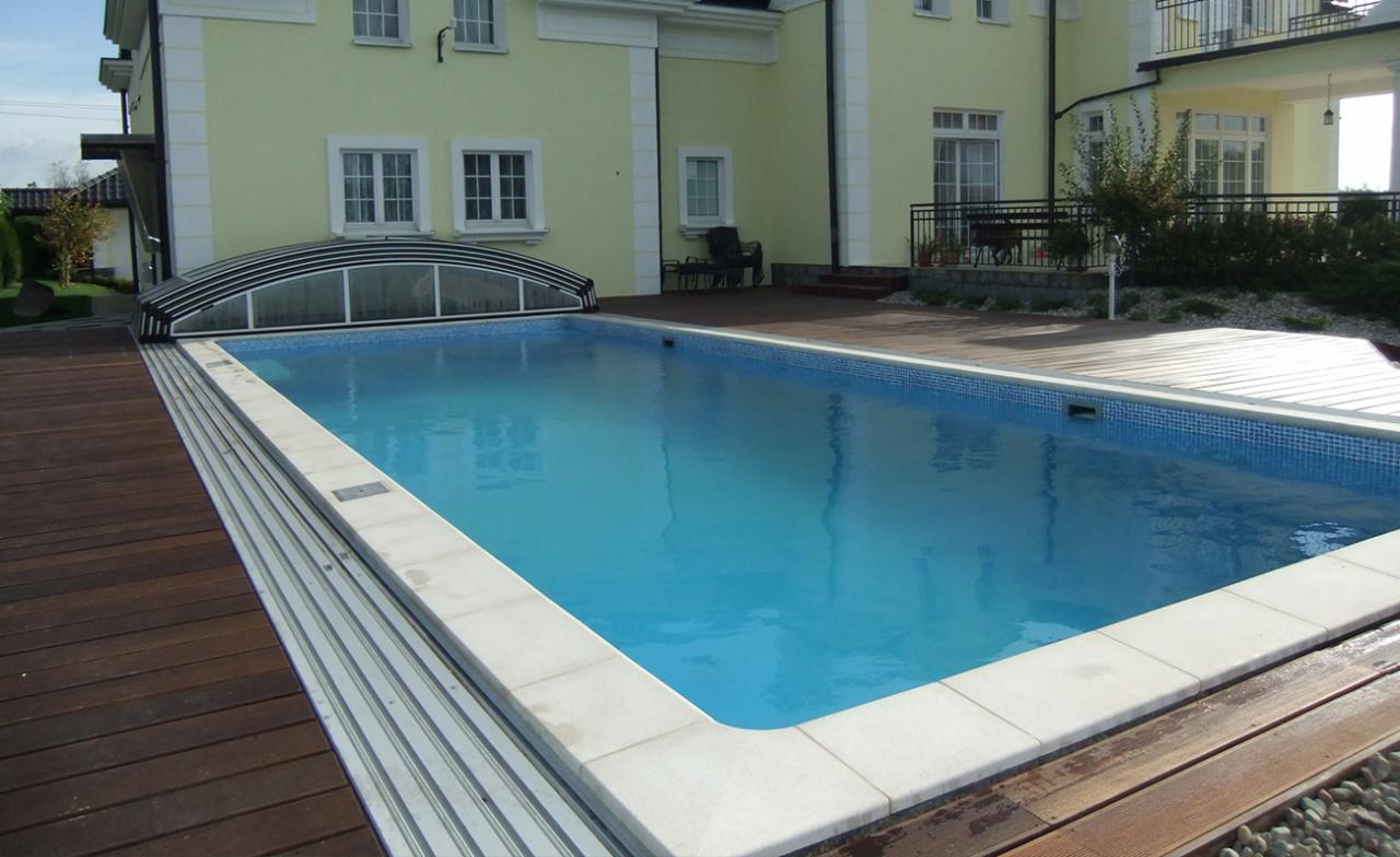 Prostokątny basen z błękitną wodą, obłożony jasnymi płytkami, z częściowo zsuniętym zadaszeniem, otoczony drewnianym tarasem, na tle pastelowożółtego domu z białymi detalami.