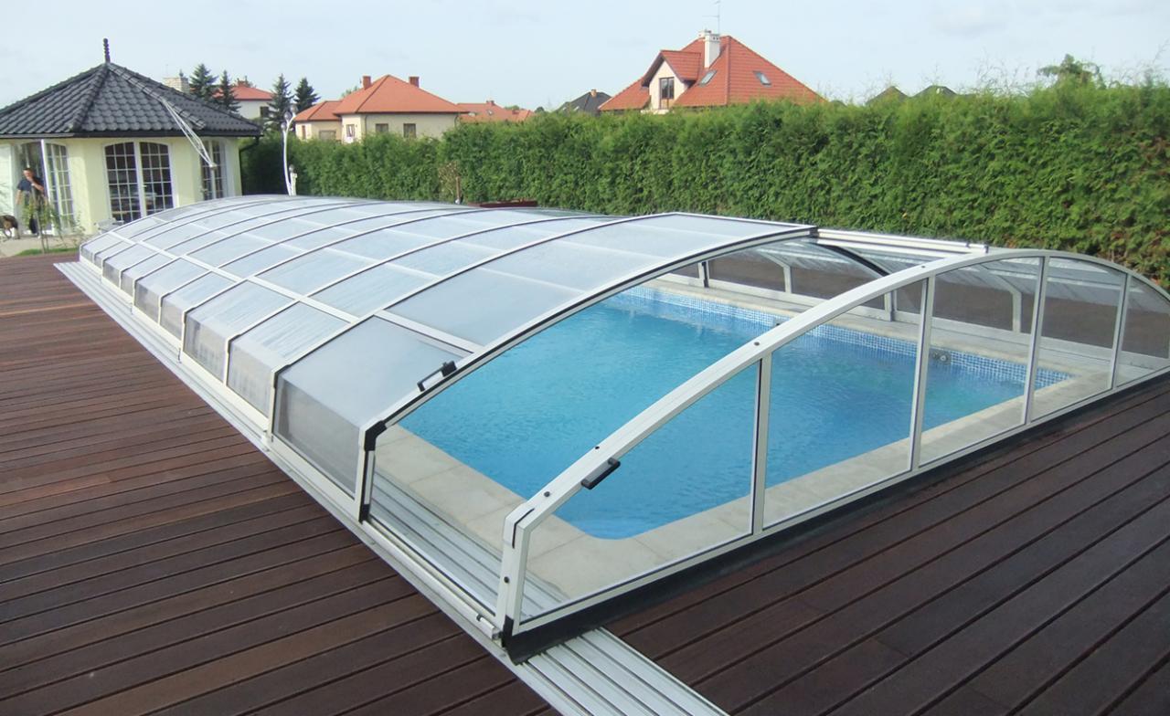 Zadaszony basen z przesuwnymi panelami na drewnianym tarasie, widok na domy jednorodzinne i żywopłot w tle.