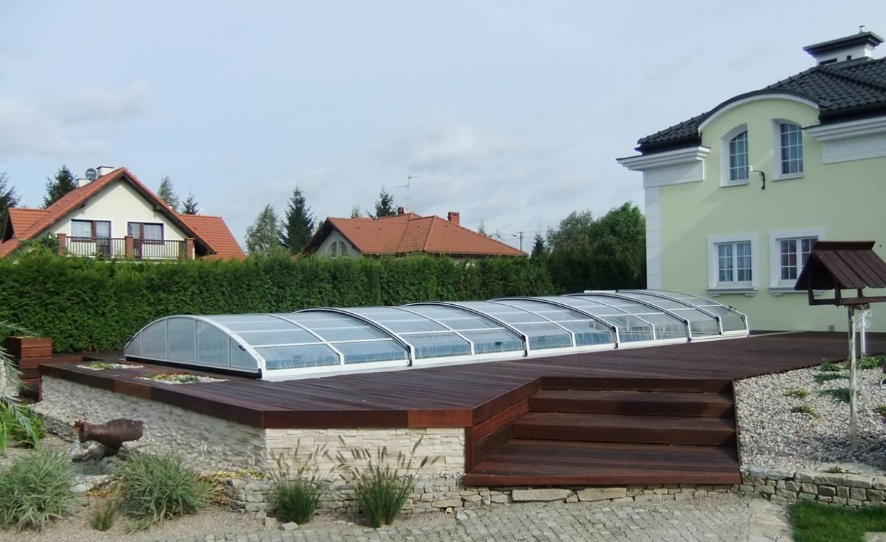 Zadaszony basen ogrodowy z drewnianym tarasem, otoczony zielenią i kamieniami, z widokiem na domy w tle.