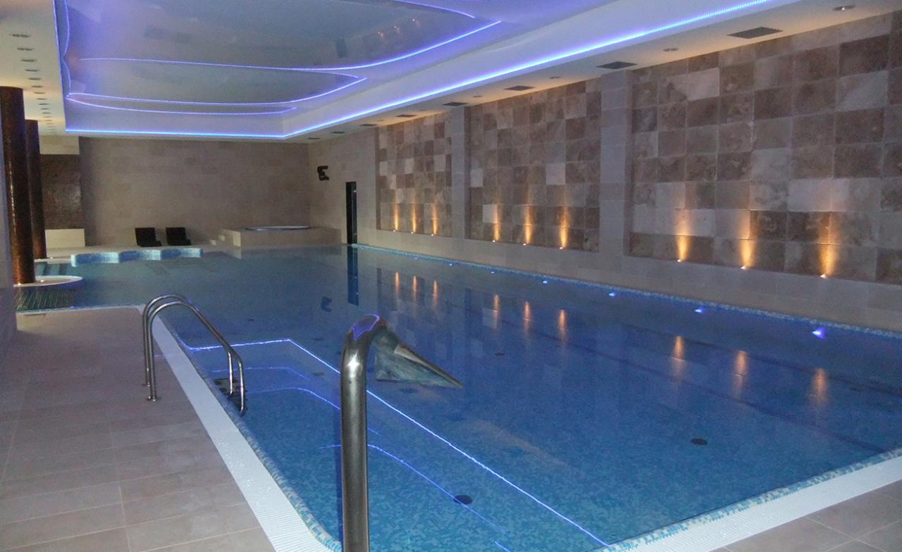 Wnętrze krytego basenu z niebieską mozaiką, oświetleniem LED i marmurową ścianą akcentującą.