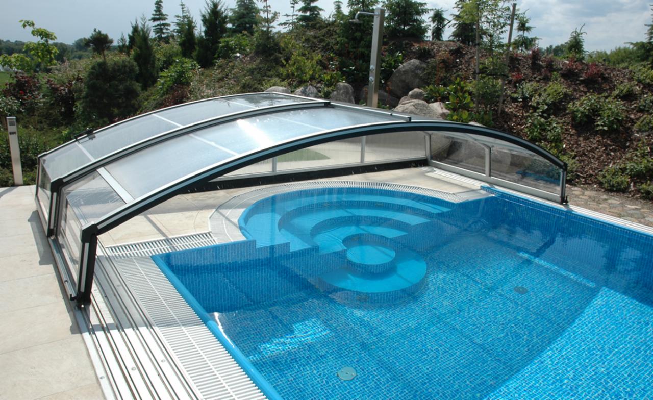 Błękitny basen z mozaiką i schodkową konstrukcją, zadaszony przesuwną konstrukcją z przezroczystego tworzywa, otoczony zielenią.