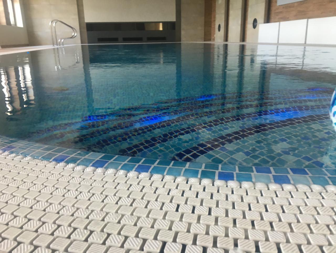 Kryty basen z mozaiką w odcieniach błękitu i białej obwódki, widoczne odbicia światła w wodzie i metalowa poręcz w tle.