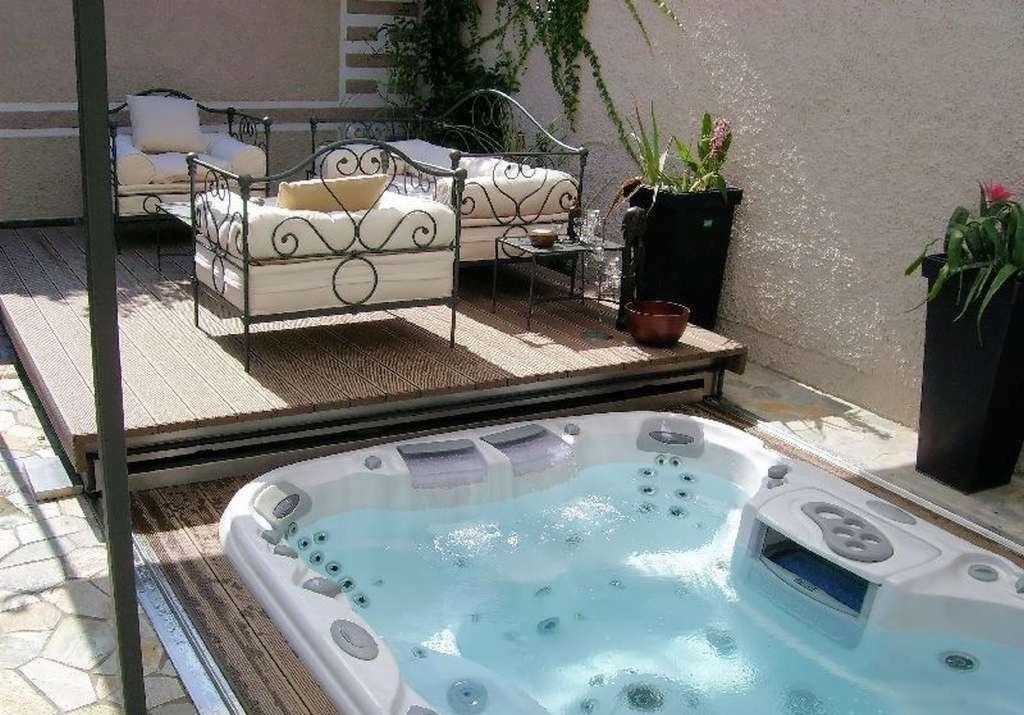 Białe jacuzzi z hydromasażem na drewnianym tarasie z metalowymi leżankami z kremowymi poduszkami w tle. Zewnętrzne otoczenie z roślinami w donicach.