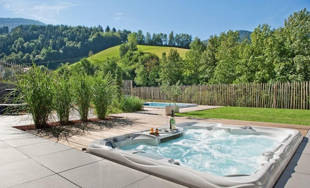 Nowoczesny, biały jacuzzi z hydromasażem na tarasie z szarych płyt, butelka wina i kieliszek na wbudowanej półce, w tle zielony trawnik, drewniany płot i drugi, mniejszy basen.