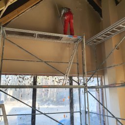 JANKOWSKI DAWID DAWBUD - Pracownik w czerwonym kombinezonie na rusztowaniu, wygładza świeżo nałożony tynk na skośnej ścianie poddasza w surowym stanie, widoczne drewniane elementy konstrukcji dachu i okno z folią ochronną.