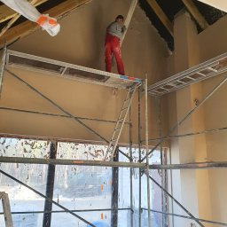 JANKOWSKI DAWID DAWBUD - Pracownik w czerwonym kombinezonie na rusztowaniu, wyrównujący tynk na poddaszu domu z odsłoniętą więźbą dachową i zabezpieczonymi oknami folią.