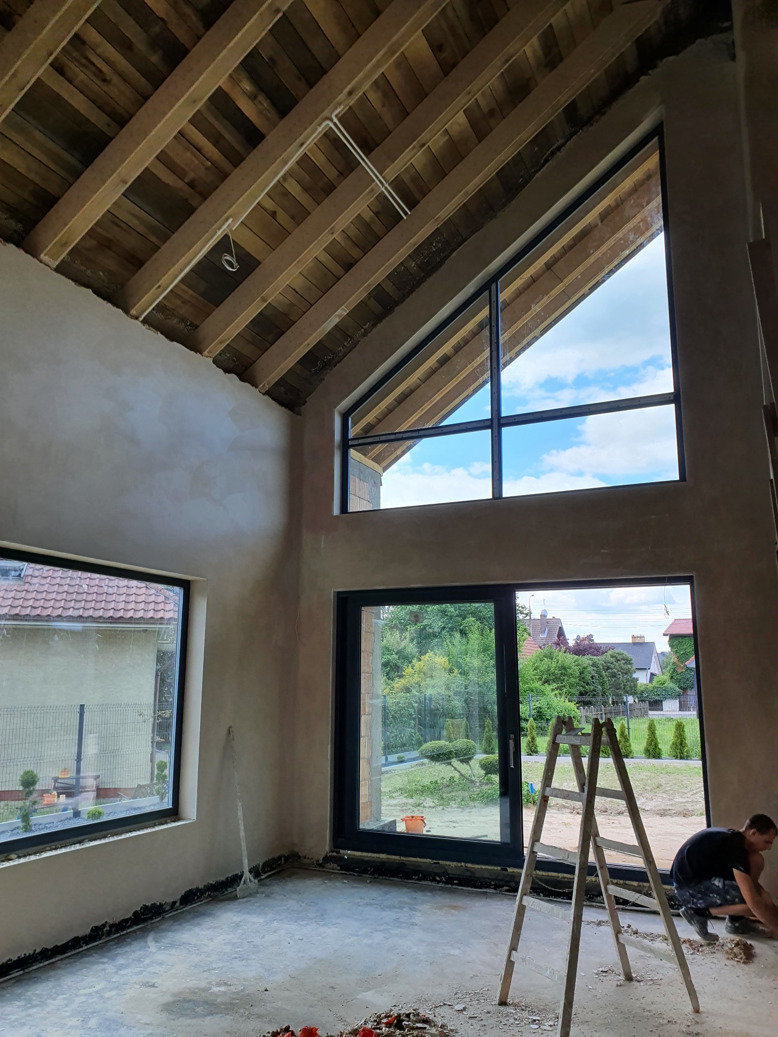 Wysokie pomieszczenie z drewnianym stropem i dużymi oknami podczas prac tynkarskich, widoczna drabina i pracownik przy ścianie.