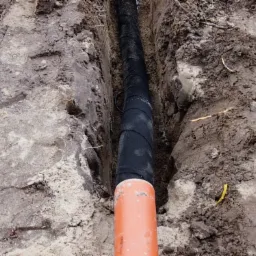 Rów w ziemi z pomarańczową rurą kanalizacyjną i czarną rurą w izolacji, widoczne ślady po wykopie.