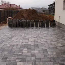 Ułożona szara kostka brukowa na posesji, w tle sterta ziemi, budynki mieszkalne i ogrodzenie z siatki.