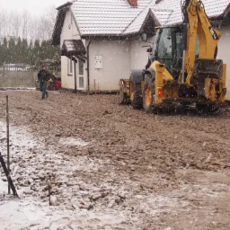 Żółta koparko-ładowarka CAT 444E przygotowuje teren pod budowę drogi w zimowej scenerii, widoczny pracownik i budynek w tle.