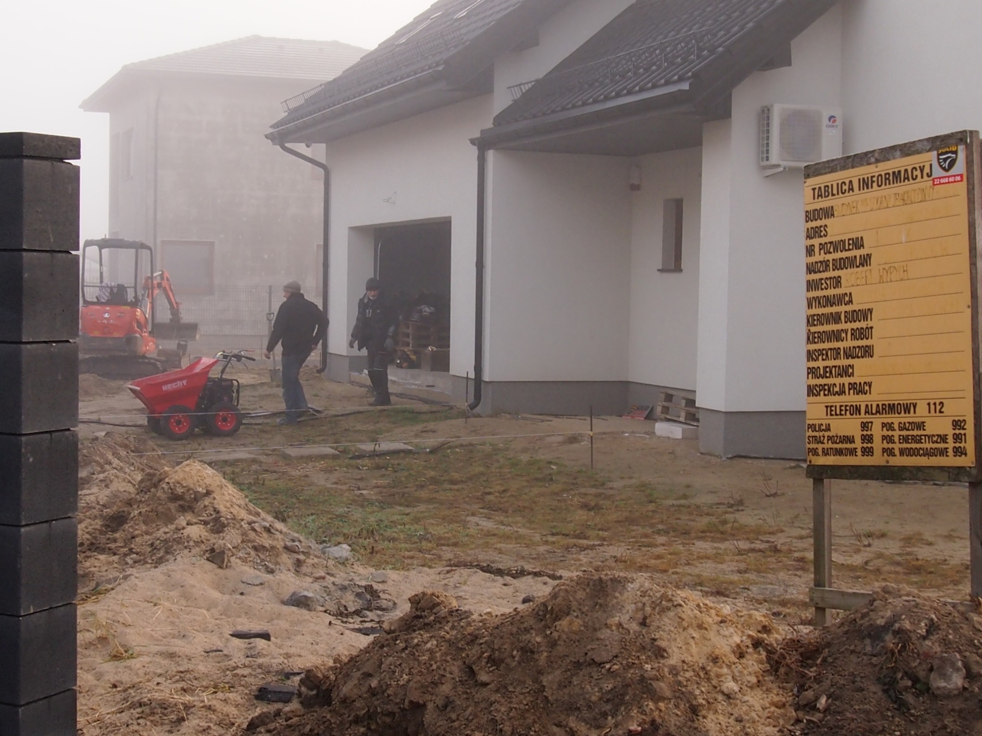 Plac budowy w mglisty dzień: widoczny nowy budynek z otwartym garażem, mini koparka, taczka motorowa, sterta ziemi i tablica informacyjna budowy z danymi inwestora i numerami alarmowymi.