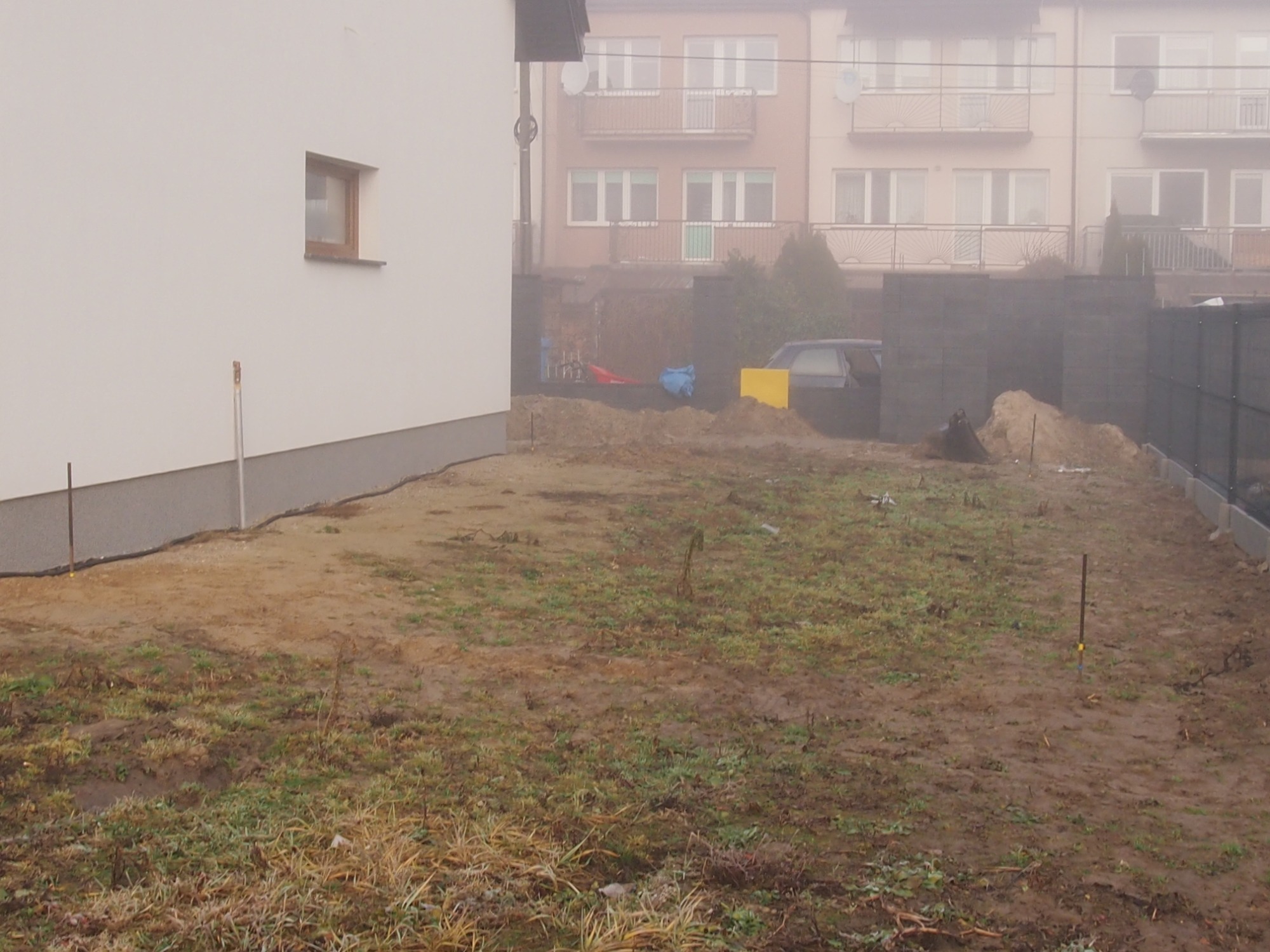 Teren przygotowany pod budowę w pochmurny dzień, widoczne wyznaczone granice i materiały budowlane na tle zabudowy mieszkalnej.