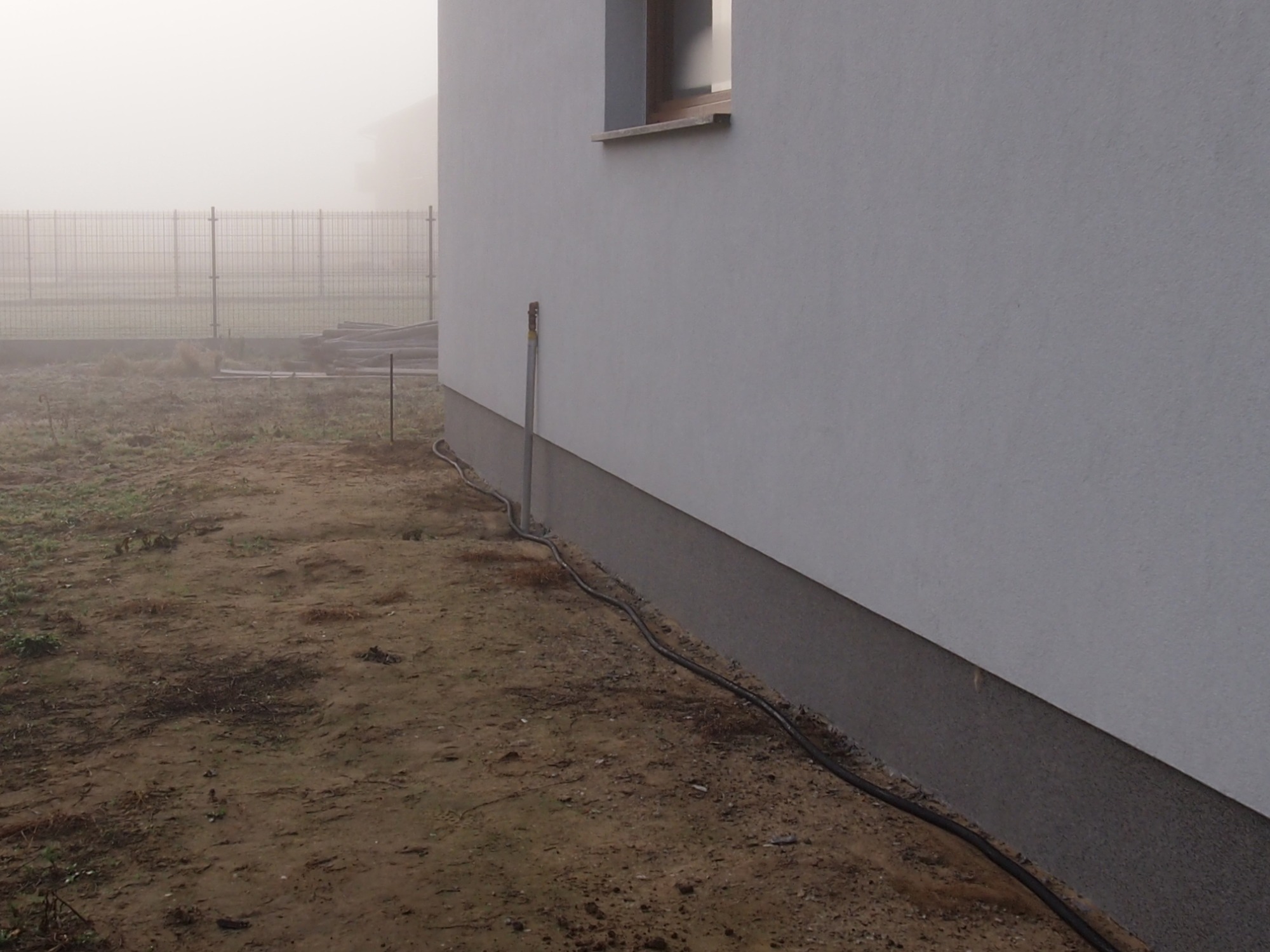 Biała ściana budynku z widoczną podmurówką i oknem, obok ziemia, w tle ogrodzenie w gęstej mgle, widoczne kable prowadzone wzdłuż ściany.