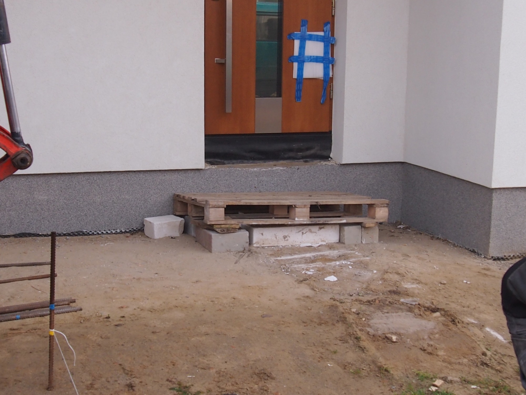Tymczasowe wejście do domu z palety i betonowych bloków, podłoże z ziemi, widoczne elementy rusztowania i fragment koparki, drzwi zabezpieczone niebieską taśmą.