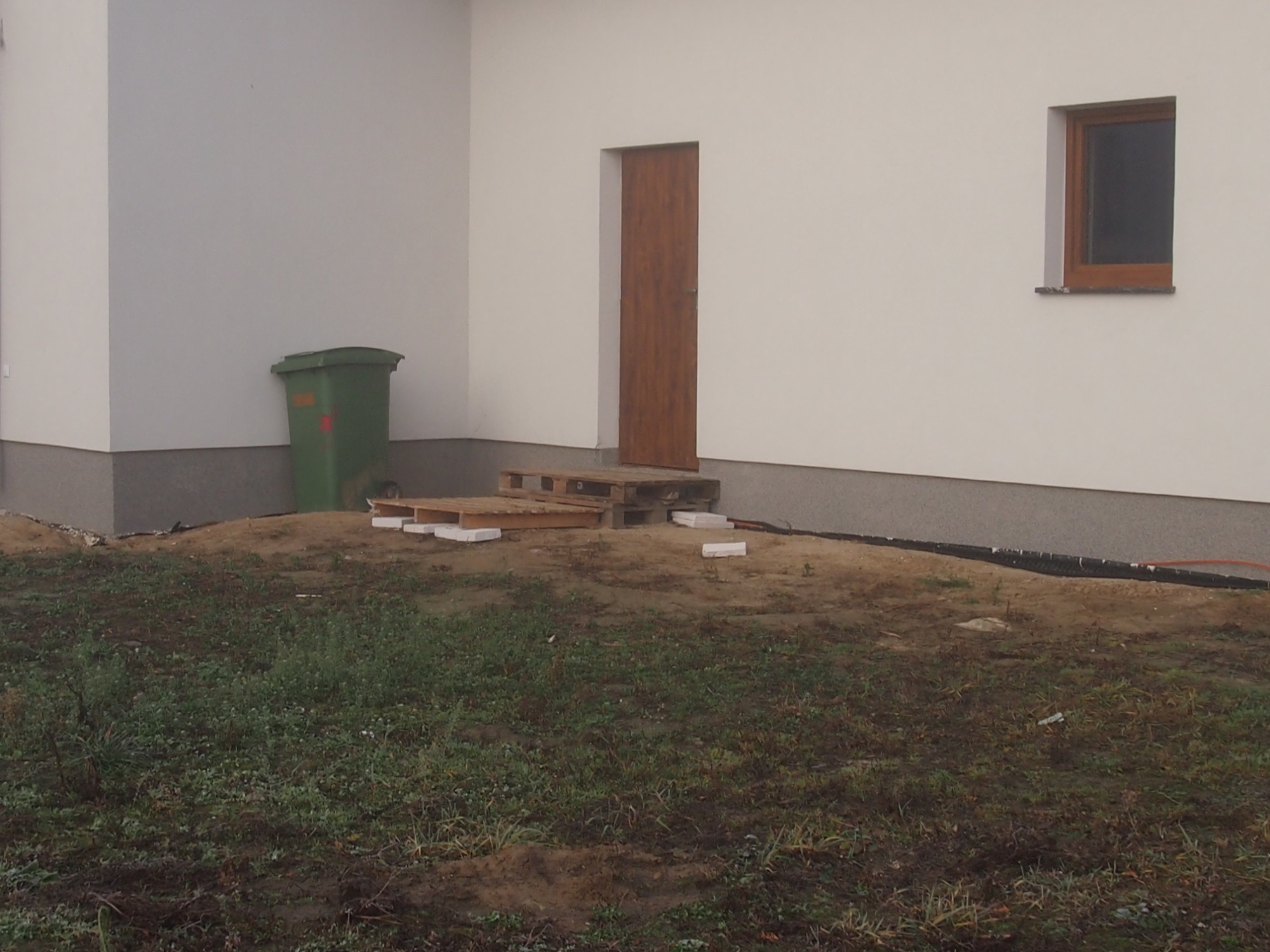 Teren przed budynkiem z przygotowanym podłożem, paletami i pojedynczymi kostkami brukowymi, widoczny śmietnik i fragment elewacji z oknem i drzwiami.