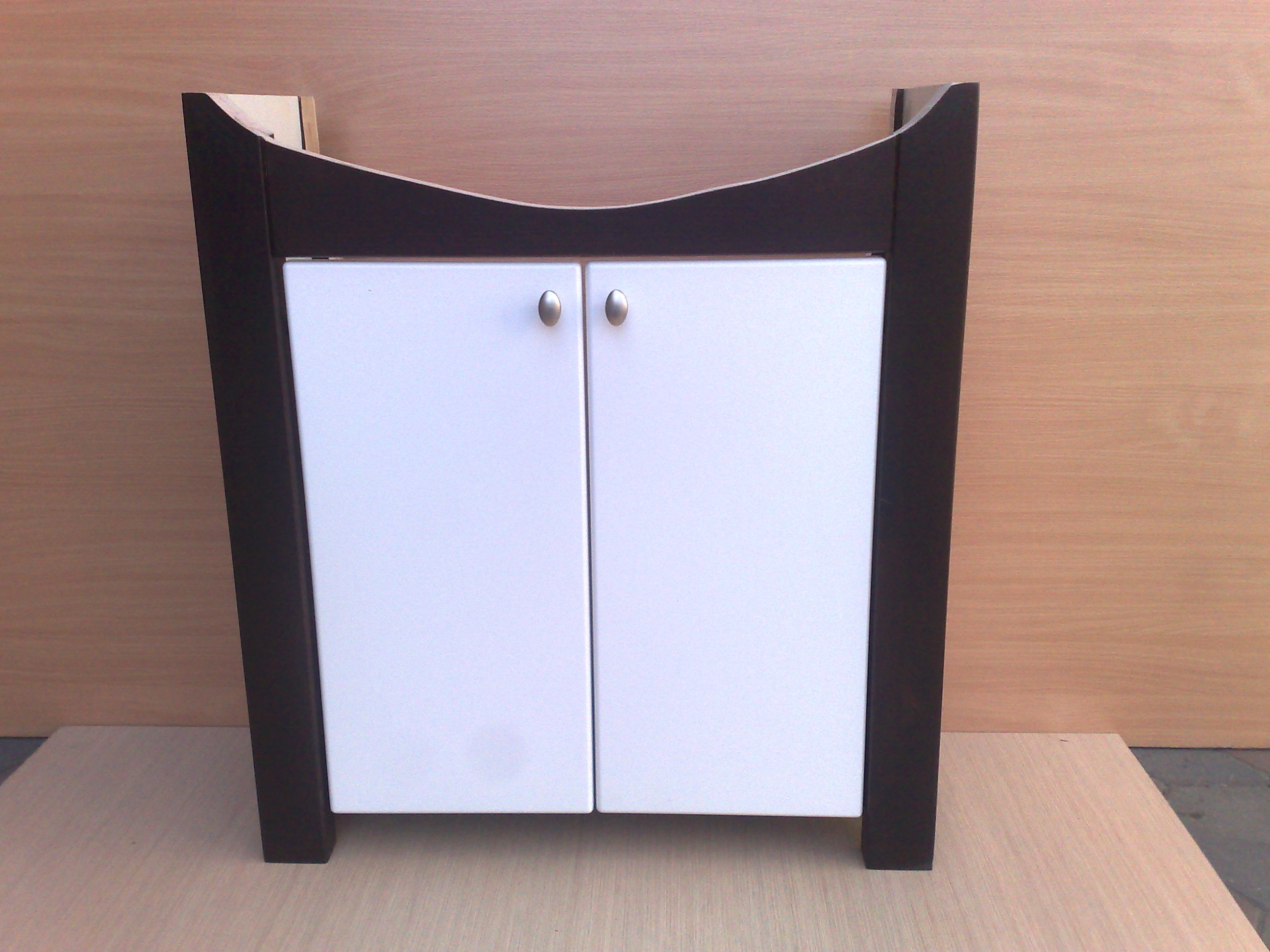 Nowoczesna szafka łazienkowa z białymi frontami i ciemnobrązową obudową, zaokrąglona u góry, z metalowymi gałkami, na tle drewnianej ściany.