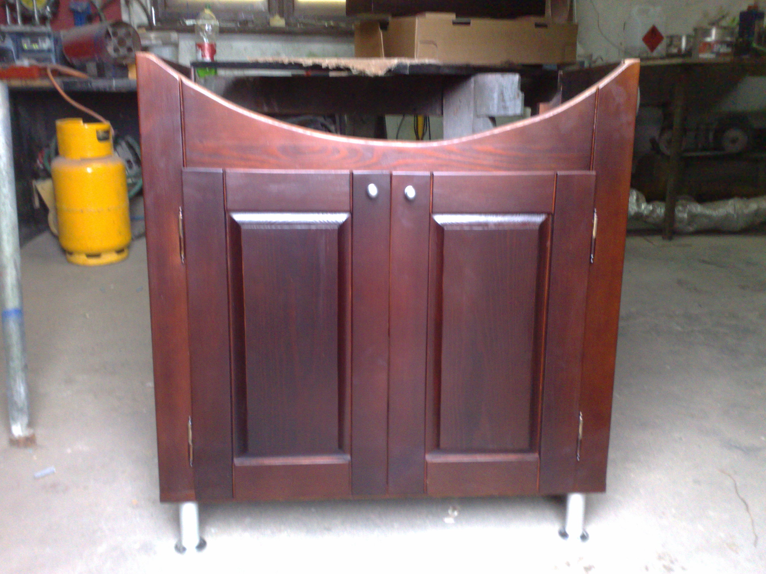 Ciemnobrązowa szafka łazienkowa z dwoma drzwiczkami i chromowanymi uchwytami, stojąca na metalowych nóżkach, sfotografowana w warsztacie, obok butla gazowa.