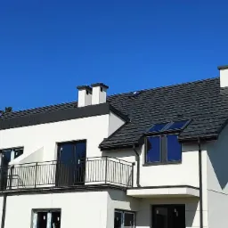 Nowoczesny dom z ciemnoszarą dachówką, białymi ścianami i czarnymi ramami okiennymi, widok z dołu na tle błękitnego nieba.