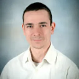 Mężczyzna w białej koszuli na tle błękitno-szarego gradientu. Portret z lekkim uśmiechem, oświetlenie z góry. Widoczne zmarszczki mimiczne.