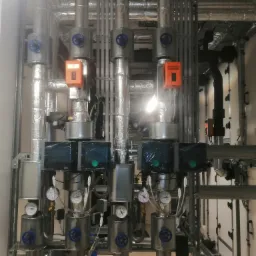Złożona instalacja rur z izolacją, zaworami z niebieskimi pokrętłami, manometrami i pomarańczowymi skrzynkami sterującymi w kotłowni lub węźle cieplnym.