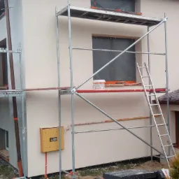 Elewacja domu w trakcie prac wykończeniowych, widoczne rusztowanie, zabezpieczone okna, wiadro z farbą i stos płyt izolacyjnych.