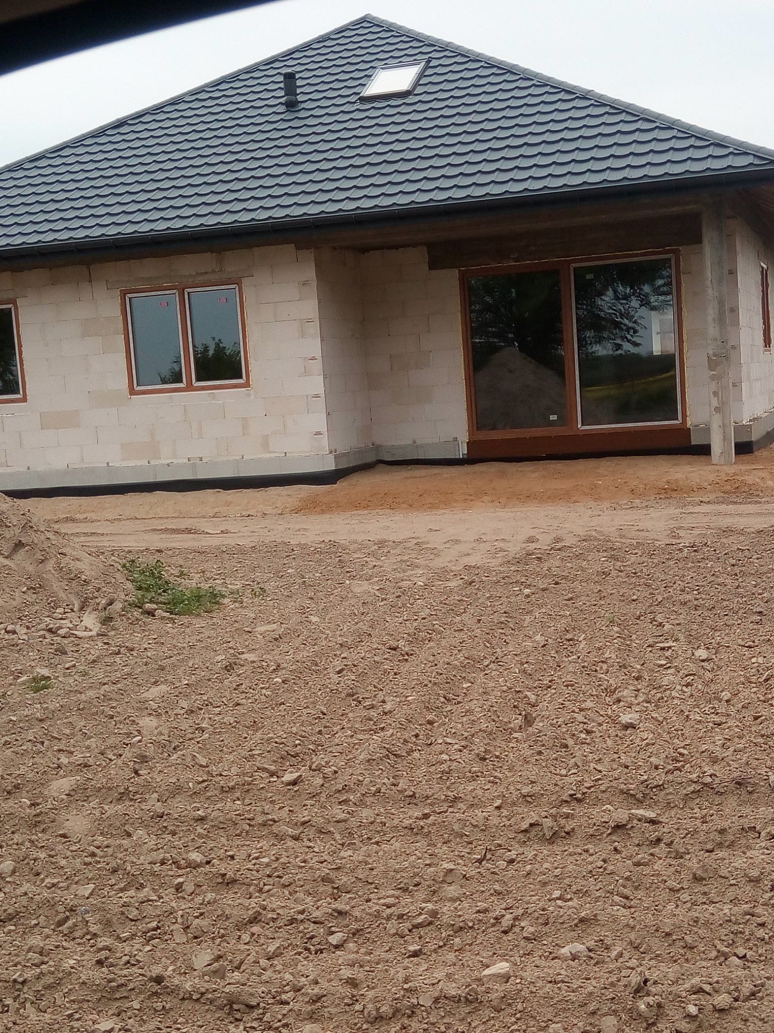 Niedokończony budynek z szarej cegły z ciemnoszarą dachówką, widoczne brązowe ramy okienne i drzwi przesuwne, teren wokół budynku niezagospodarowany, z ziemią i gruzem.