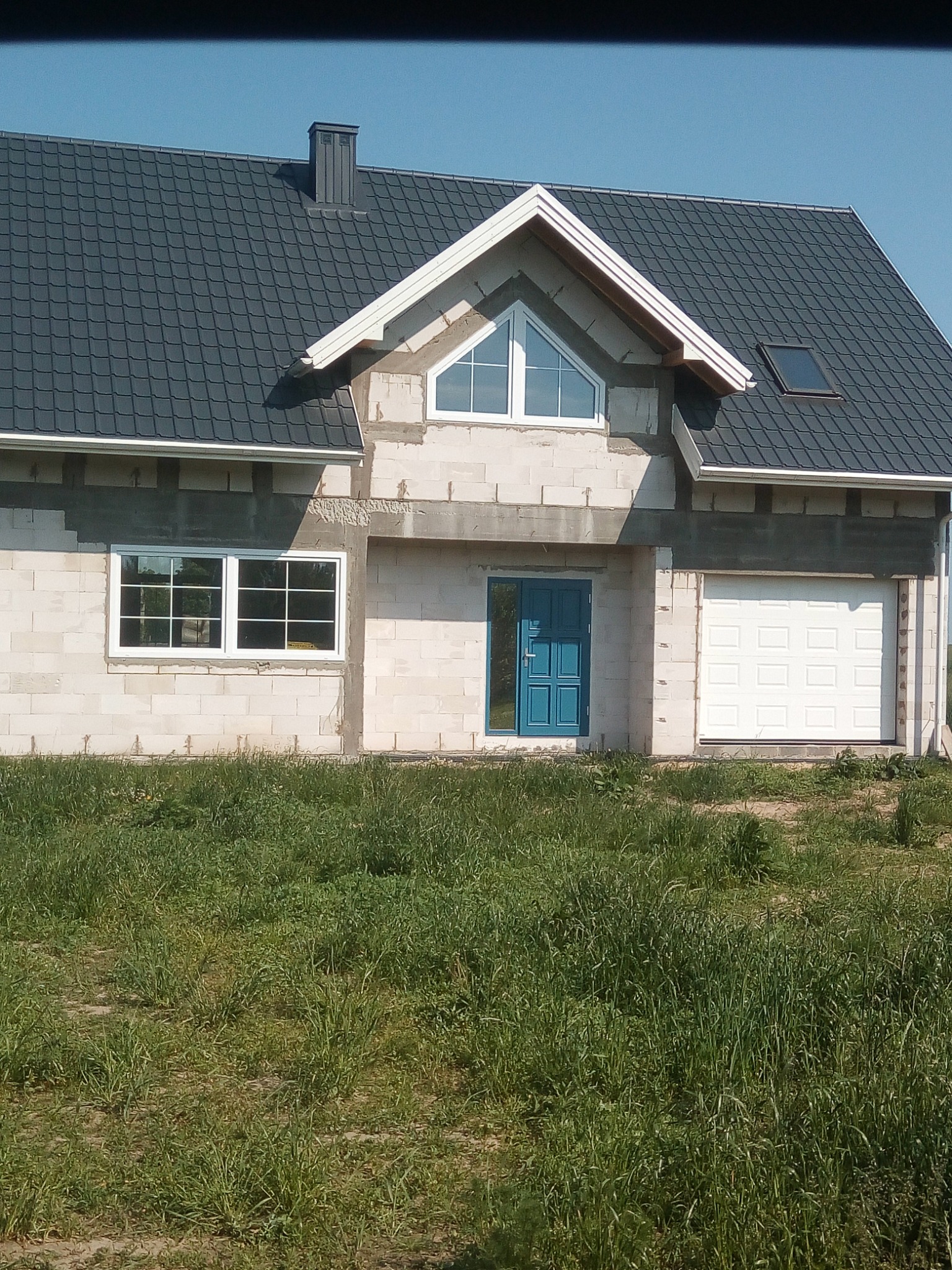 Surowy dom murowany w trakcie budowy z szarą dachówką, niebieskimi drzwiami wejściowymi i białymi oknami, widok na elewację frontową z trawiastym terenem.