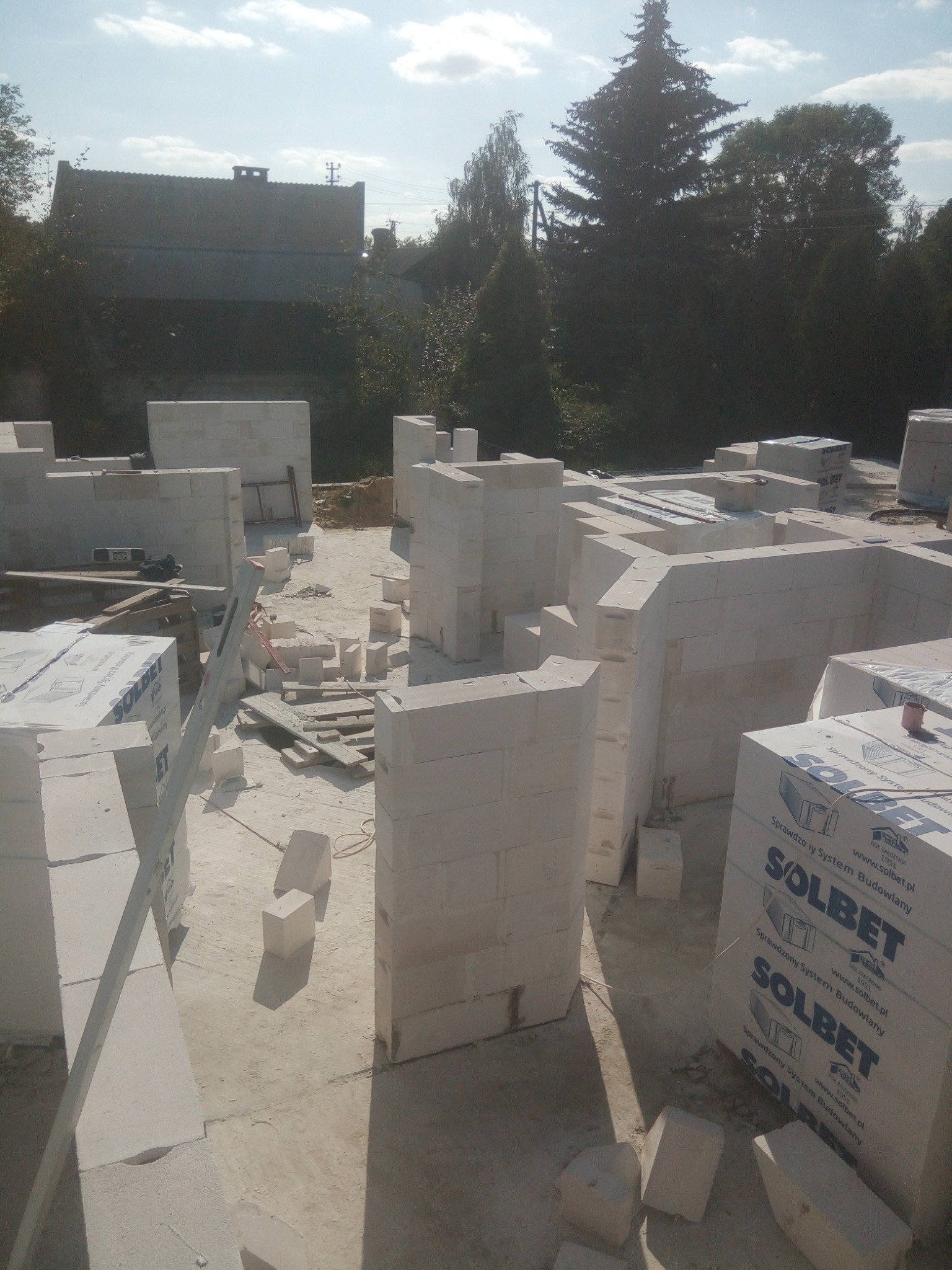 Wznoszenie ścian parteru z białych bloczków betonu komórkowego Solbet. Widoczne palety z materiałem budowlanym, poziomica i fragment krajobrazu z zabudowaniami w tle.