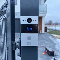 FlexiSystem - Zamontowany wideofon Akuvox na słupku bramy posesji. Widoczny panel z kamerą, podświetlony przycisk dzwonka i elementy montażowe. W tle dom jednorodzinny.