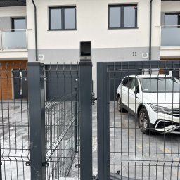 FlexiSystem - Nowoczesna posesja z automatyczną bramą wjazdową, widoczny napęd bramy. W tle biały samochód osobowy i budynek z szarą elewacją. Brama z metalowej siatki.