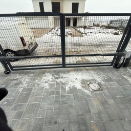 FlexiSystem - Automatyczna brama posesji z siatki, widok z bliska na mechanizm otwierający. W tle biały budynek i częściowo widoczny biały van. Kostka brukowa na pierwszym planie.