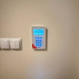 FlexiSystem - Biały panel alarmowy z niebieskim wyświetlaczem zamontowany na ścianie obok włącznika światła. Widoczny aktualny czas i data.