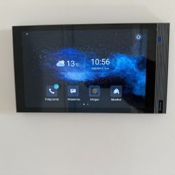 FlexiSystem - Nowoczesny panel dotykowy wideodomofonu Akuvox z interfejsem użytkownika, zamontowany na białej ścianie. Widoczny ekran z ikonami połączeń i wiadomości.