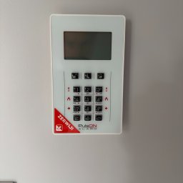FlexiSystem - Panel alarmowy PulsON z ekranem LCD i klawiaturą numeryczną, zamontowany na szarej ścianie. Widoczny czerwony element z napisem 'ZERWIJ!'.