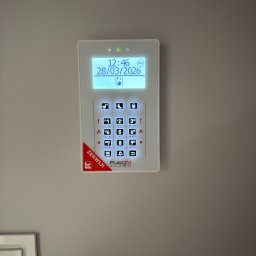 FlexiSystem - Panel alarmowy Pulson Alarm na ścianie, z podświetlanym ekranem LCD pokazującym datę i godzinę. Widoczne przyciski numeryczne i funkcyjne. Czerwony napis 'ZERWIJ!' w lewym dolnym rogu.
