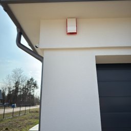 FlexiSystem - Zewnętrzna ściana domu z zamontowaną sygnalizacją alarmową, widoczna kamera monitoringu pod dachem i fragment bramy garażowej. Ujęcie z dołu, w tle drzewa.