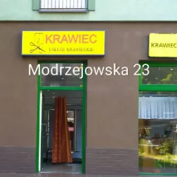 Fasada budynku z szyldem 'Krawiec' i adresem Modrzejowska 23, widoczne okna i wejście do zakładu krawieckiego.