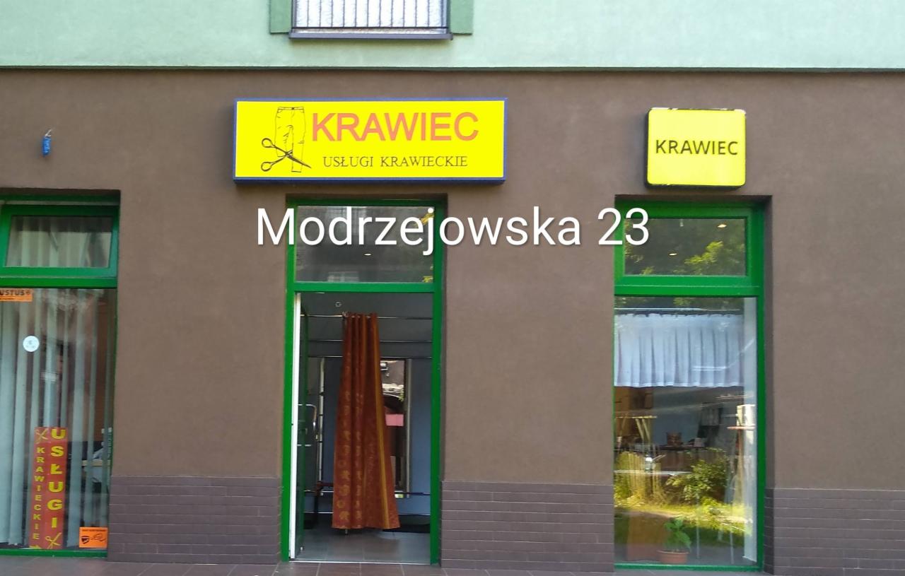 Fasada budynku z szyldem 'Krawiec' i adresem Modrzejowska 23, widoczne okna i wejście do zakładu krawieckiego.