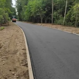 Świeżo wyasfaltowana droga z krawężnikiem, widoczny walec drogowy w oddali, otoczona zielenią drzew.